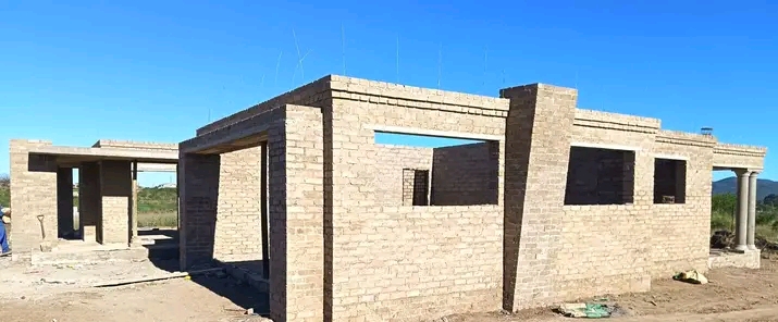 Single-story brick house under construction with classical columns and open doorways, rural setting with mountain views